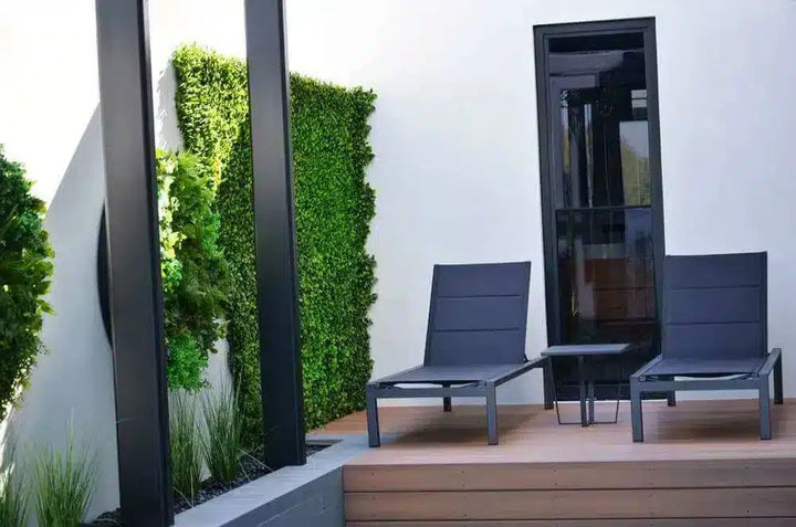 Two modern dark gray outdoor lounge chairs with a small matching side table on a wooden deck, set against a white wall with a black-framed glass door and a green vertical garden.