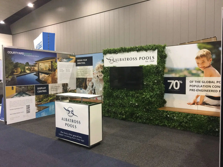 An exhibition booth for Albatross Pools, featuring a white and navy branded display table, large promotional wall panels with pool imagery, and a green foliage screen backdrop.