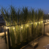 Premium 180cm potted artificial Yucca/Reed Grass lining a balcony at dusk, illuminated by warm string lights and creating a lush green privacy screen.