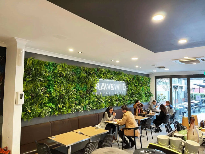 A restaurant interior with a large green living wall featuring the 'RAW & WILD MARKETPLACE' sign. Several people are seated at tables in a modern dining area with a dark ceiling and large windows.