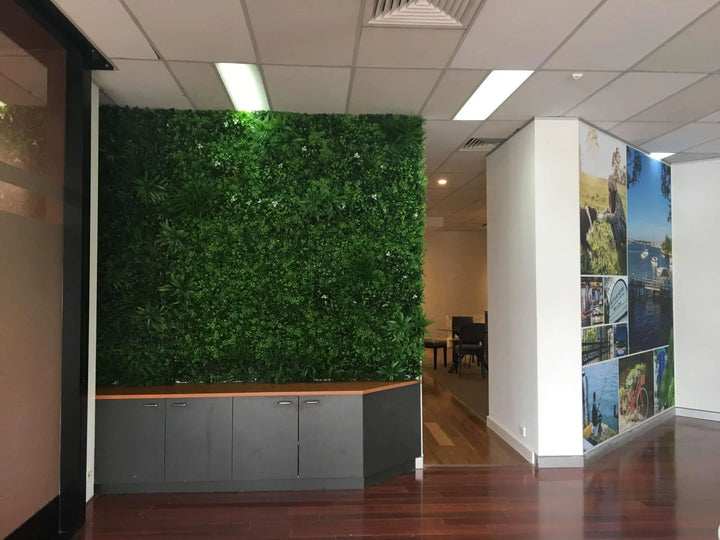 An indoor office or lobby space with a large artificial green wall mounted above gray storage cabinets, alongside a white wall with vertical framed landscape photographs.