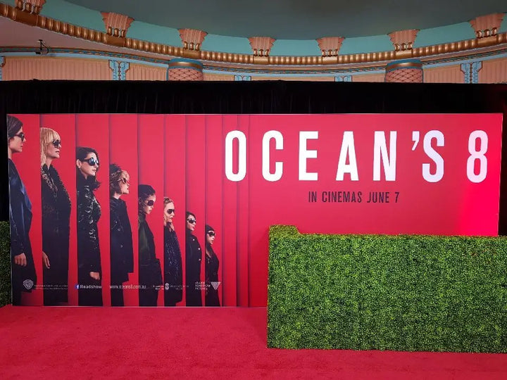A movie event backdrop for Ocean's 8 featuring a large red panel with the film title in white, character silhouettes, and release text, set on a red carpet with a green hedge in front.