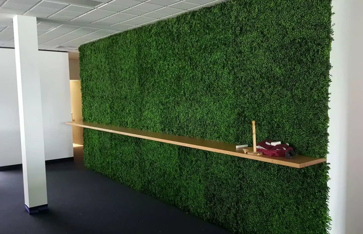 A large artificial green foliage wall mounted on an office wall, with a long light wood floating shelf mounted below it. The room has a white ceiling, white structural columns, and a dark floor.