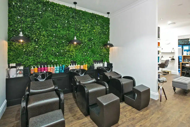 A modern salon interior with multiple dark leather shampoo chairs arranged in front of a large green foliage wall, wood flooring, pendant lights, and a counter displaying haircare bottles.