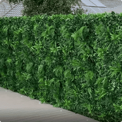 A dense green artificial hedge wall made of layered faux leaves and palm-like foliage, shown outdoors with a paved surface in front and part of a roof visible above.