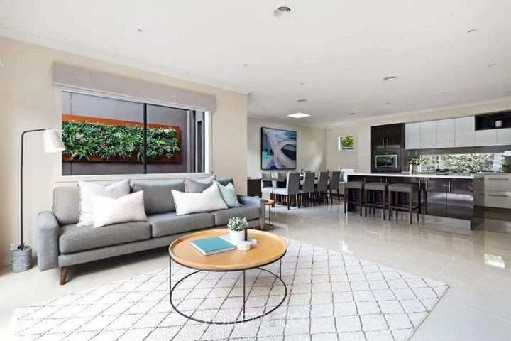 A modern open-plan living room with a gray sofa, round wooden coffee table, white patterned rug, and a large kitchen area in the background.