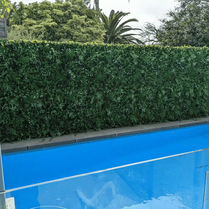 A tall, dense green artificial hedge running parallel to a bright blue swimming pool, with trees and foliage visible in the background.