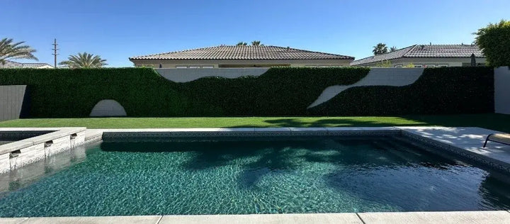 A backyard swimming pool with clear blue water in the foreground, a tall green hedge, manicured lawn, and residential house with a tiled roof under a bright blue sky.