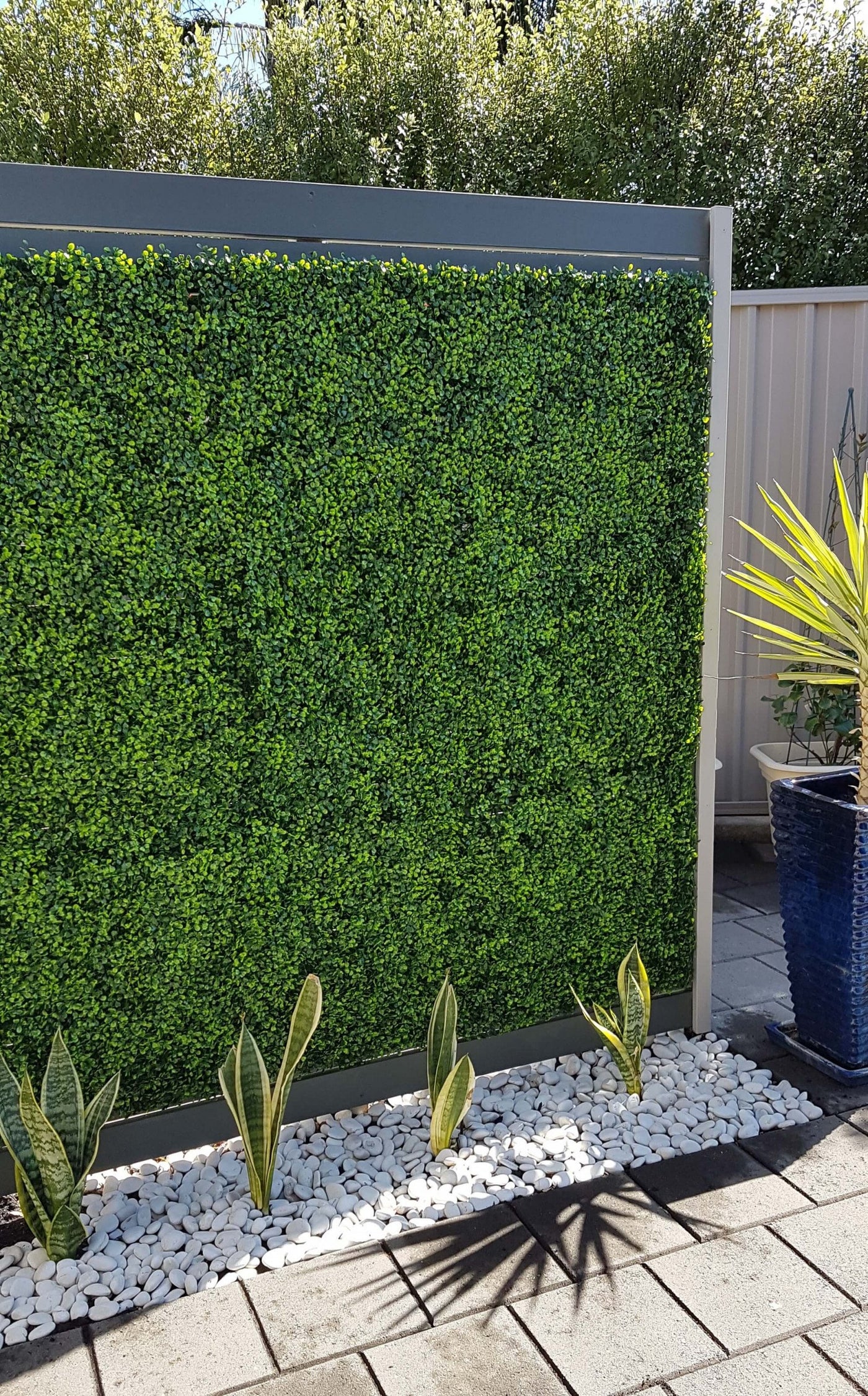 Artificial green wall panel in a garden setting with plants and pebbles.