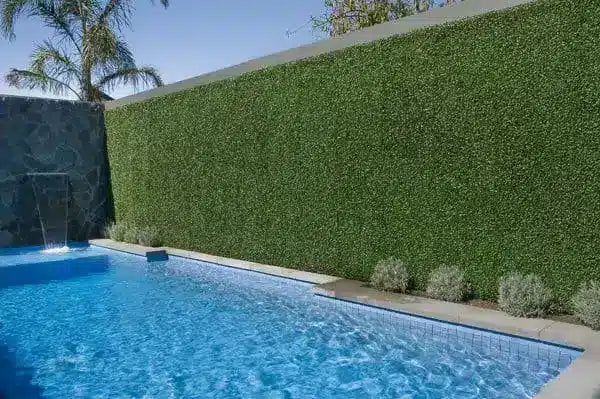 A tall, dense green artificial hedge wall running alongside a blue swimming pool, with a stone waterfall feature on the left and small shrubs along the base.