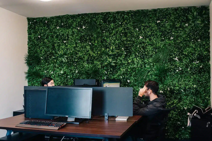 A modern office space featuring a large green artificial foliage wall behind two people seated at a wooden desk with computer monitors.