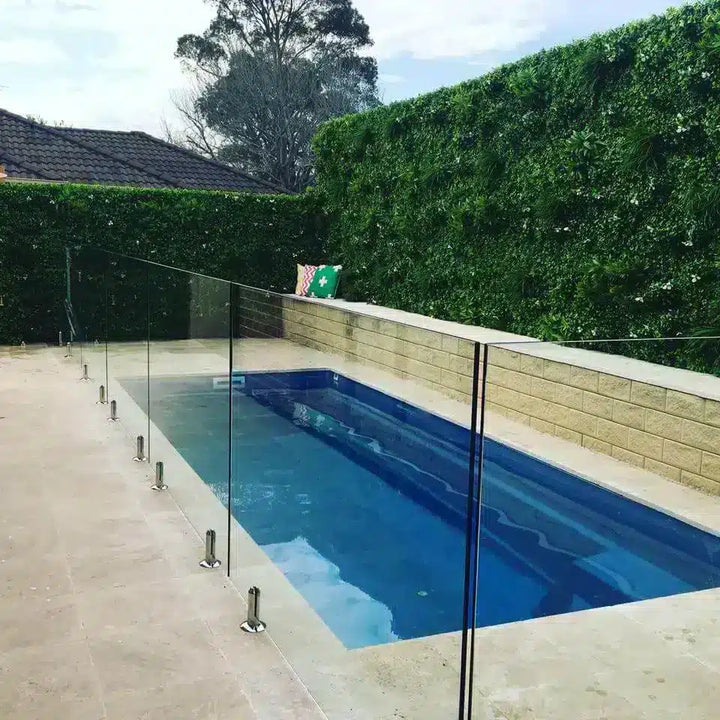 A modern rectangular swimming pool with blue water and a clear glass safety fence supported by stainless steel posts, set in a backyard with a stone wall, green hedge, and tiled patio.