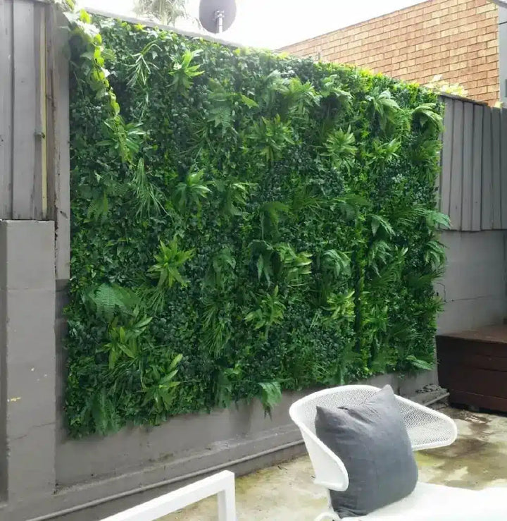 A large artificial green living wall made of dense faux foliage panels mounted on a gray concrete wall in an outdoor patio setting, with a white chair and gray cushion in the foreground.