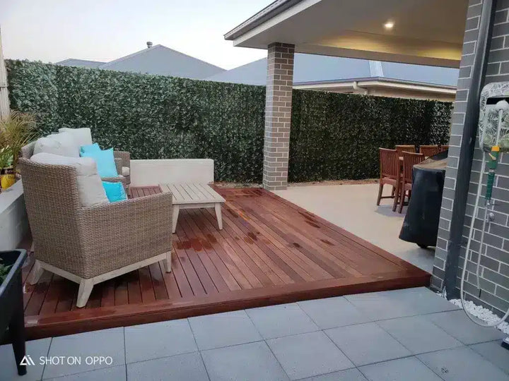 A raised wooden deck with a reddish-brown finish in a backyard patio setting, featuring a wicker armchair with white and blue cushions, a white bench, and a brick pillar supporting a covered seating area.
