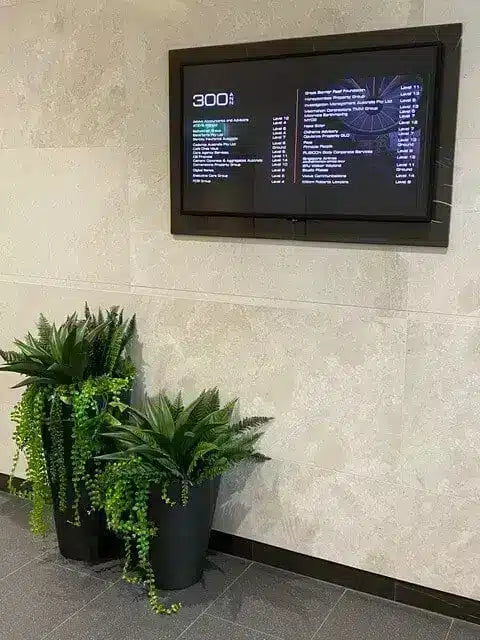 A black digital display screen mounted on a light beige textured wall, showing a list of items or menu information. Below the screen are two black planters with green artificial foliage and trailing vines, set against a gray tiled floor.