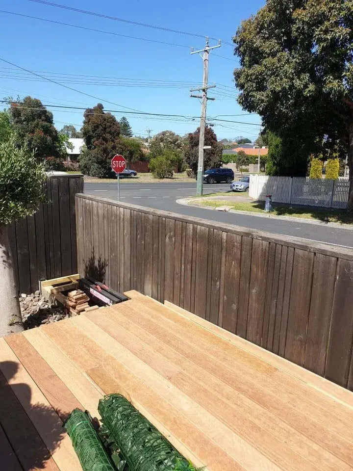 A large sheet of light-colored wood planks on a deck, with a dark wooden fence, a green roll of material, and an outdoor suburban street scene in the background under a blue sky.