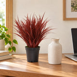 Burgundy artificial grass in its black pot beside a ceramic vase on a timber desk.