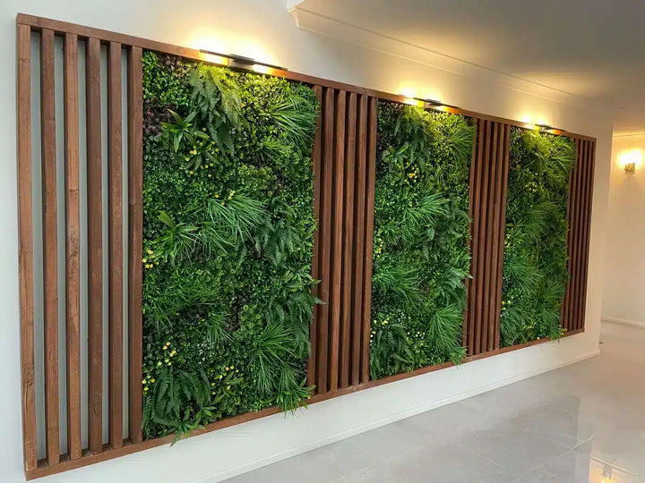 A large indoor vertical green wall installation with multiple rectangular panels of dense green foliage separated by vertical wooden slats, mounted on a white wall with warm wall lights above.
