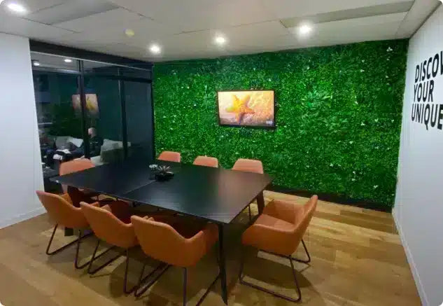 A modern conference room featuring a black rectangular meeting table surrounded by eight orange upholstered chairs, set against a prominent green artificial foliage wall with a mounted TV.