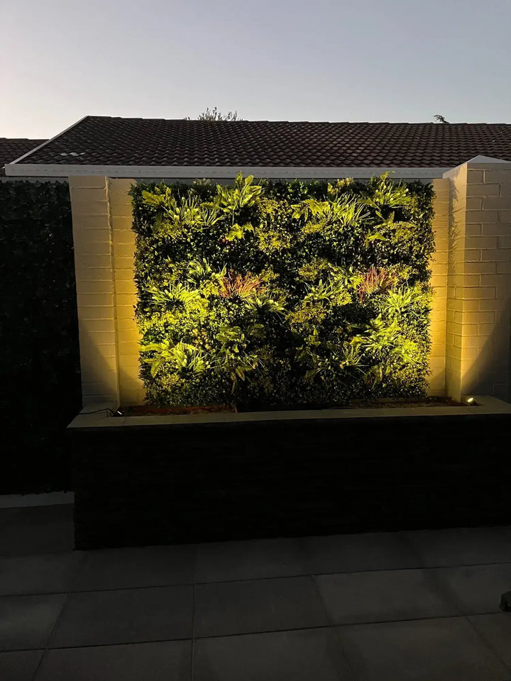 A vertical living wall garden illuminated by warm yellow LED lights, set between two light beige brick pillars under a tiled roofline.