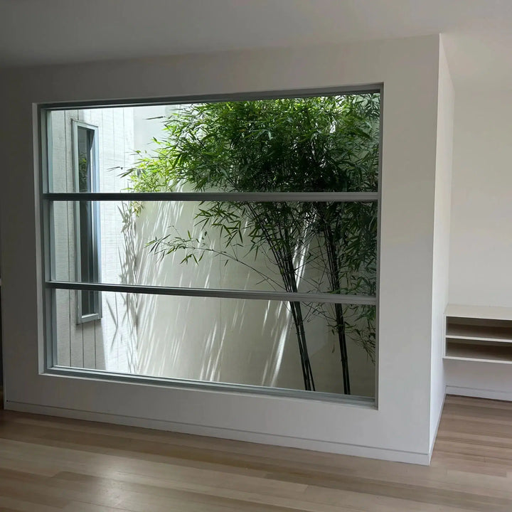 A large modern floor-to-ceiling window with three horizontal metal bars, set into a white wall in a minimalist room with light wood flooring. A green bamboo plant is visible outside.