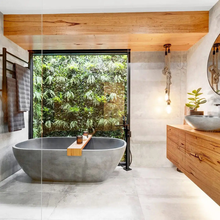 A modern freestanding concrete bathtub in a minimalist bathroom with a large glass door opening to a lush green garden, a wooden vanity with a stone sink, and warm wood ceiling accents.
