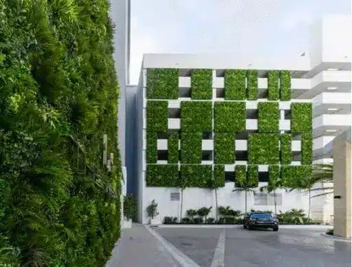 A modern white building facade covered in a grid pattern of green climbing plants, with a tall dense vertical green wall on the left and a paved driveway in front.