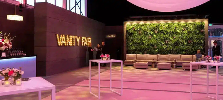 Vanity Fair event space with a large illuminated 'VANITY FAIR' sign on a dark wall, a green living wall, pink ambient lighting, and multiple white tables and seating areas.