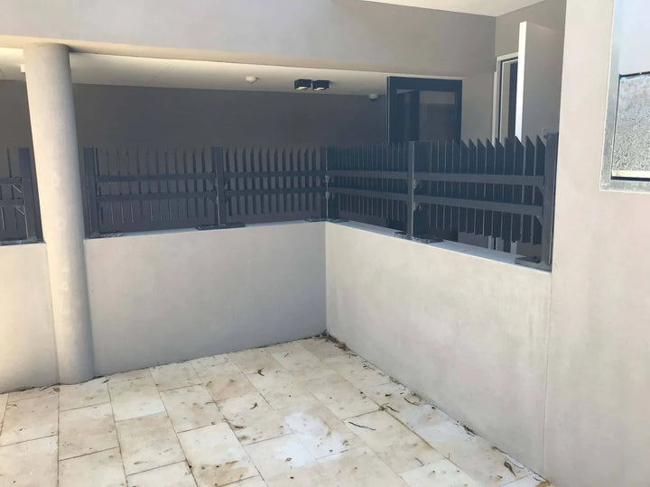 An outdoor patio or courtyard area with light beige tiled flooring, low beige walls, a dark gray metal fence, and a covered walkway overhead.