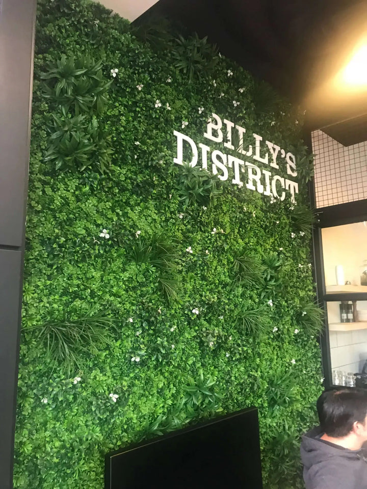 A green artificial foliage wall with the words 'BILLY'S DISTRICT' in white lettering, shown indoors with part of a person visible at the lower right.