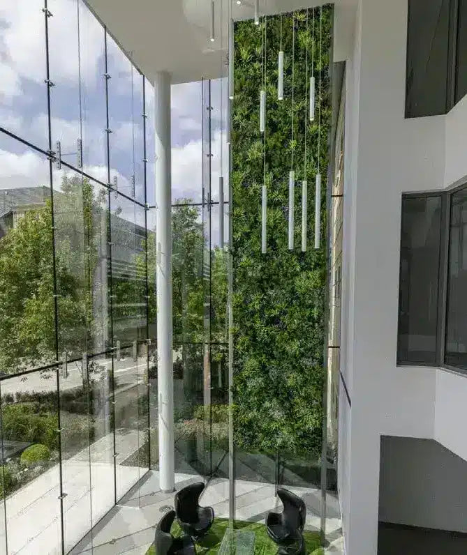 A modern interior architectural scene with a tall vertical green wall, large glass windows, white columns, hanging light tubes, and two black lounge chairs on a green floor.