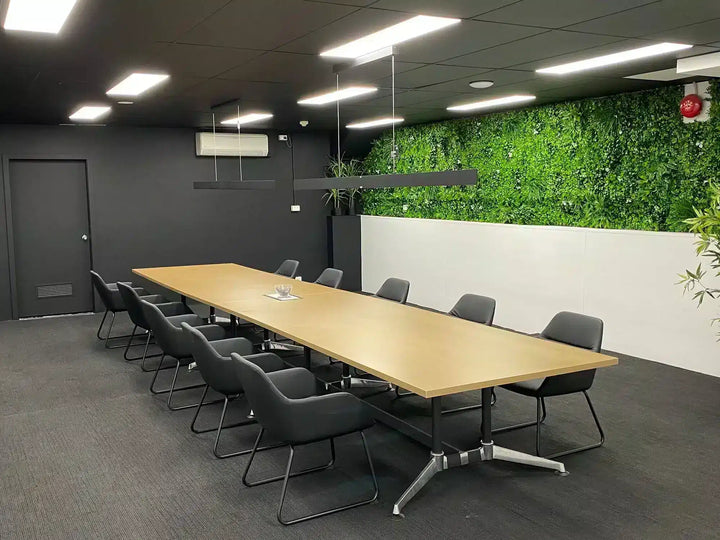 A modern conference room with a long rectangular light wood table surrounded by black office chairs, set against dark gray walls and a prominent green living wall.