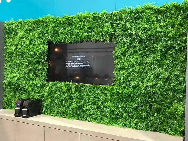 A green artificial foliage wall with a central black screen displaying text, mounted above a beige counter with two black coffee machines in the foreground.