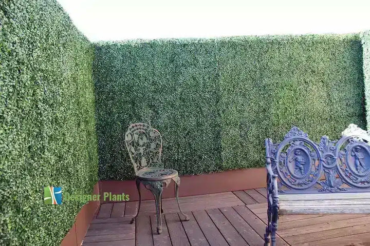 A decorative artificial hedge wall on a wooden deck, styled with a metal garden chair and part of a bench. A 'Hedge Plants' logo watermark is visible in the lower-left corner.