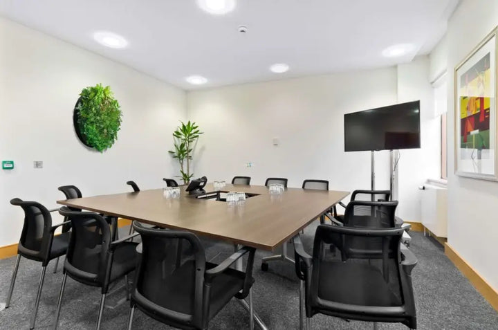 A modern conference room with a rectangular wooden table surrounded by black mesh office chairs, a wall-mounted flat screen TV, a potted plant, and a green circular wall decor piece.