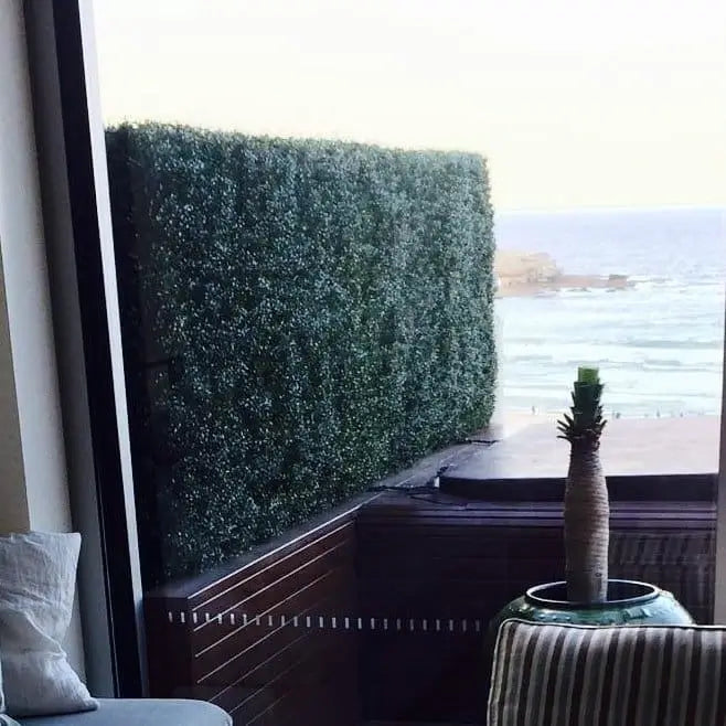 A tall, dense artificial green hedge wall installed on a dark wooden balcony overlooking the ocean.
