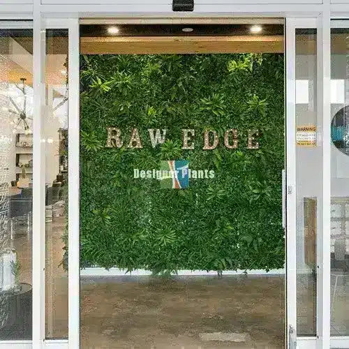 A large green artificial foliage wall with the words 'RAW EDGE' and 'Designer Plants' displayed on it, situated behind glass doors in a storefront setting.