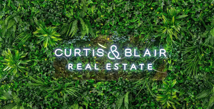 A green foliage wall with the text 'CURTIS & BLAIR REAL ESTATE' centered in white neon-style lettering.