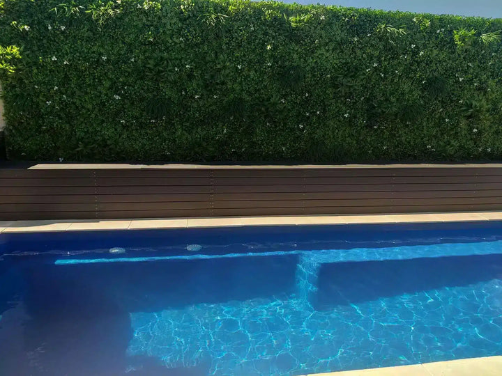 A swimming pool with bright blue water in the foreground, a dark brown wooden deck, and a tall green hedge in the background.