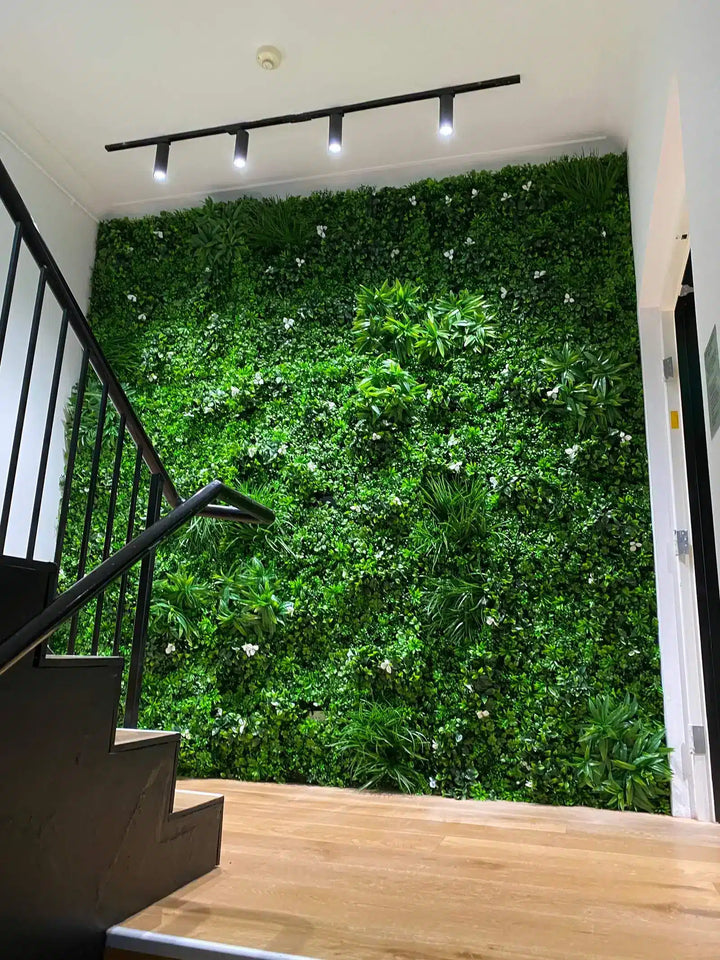 A large vertical green wall made of dense artificial foliage stands behind a wooden staircase landing, with a black track light mounted on the ceiling above.