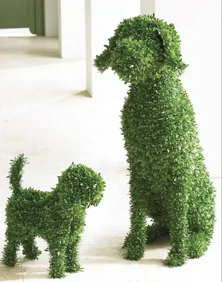 Two green topiary dog sculptures, one large and one small, made from dense leafy greenery, standing on a light-colored floor in an indoor setting.