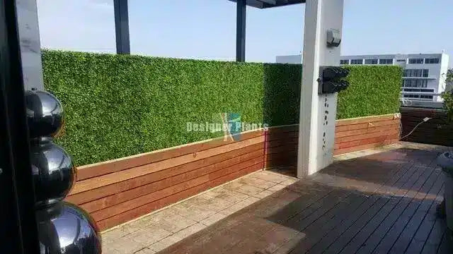 A green artificial hedge wall mounted on a wooden planter box on a modern outdoor terrace with a wooden deck and white structural columns.