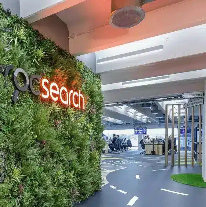A modern office interior with a large green living wall on the left featuring a glowing orange 'rqc search' logo and a magnifying glass icon. The floor has a curved white line resembling a road, and people are seated in a meeting area in the background under bright ceiling lights.